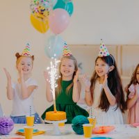 Overjoyed children friends clap hands and looks at big sparkle on cake, stand near festive table with present, cups and holiday attributes, have fun together, celebrate birthday